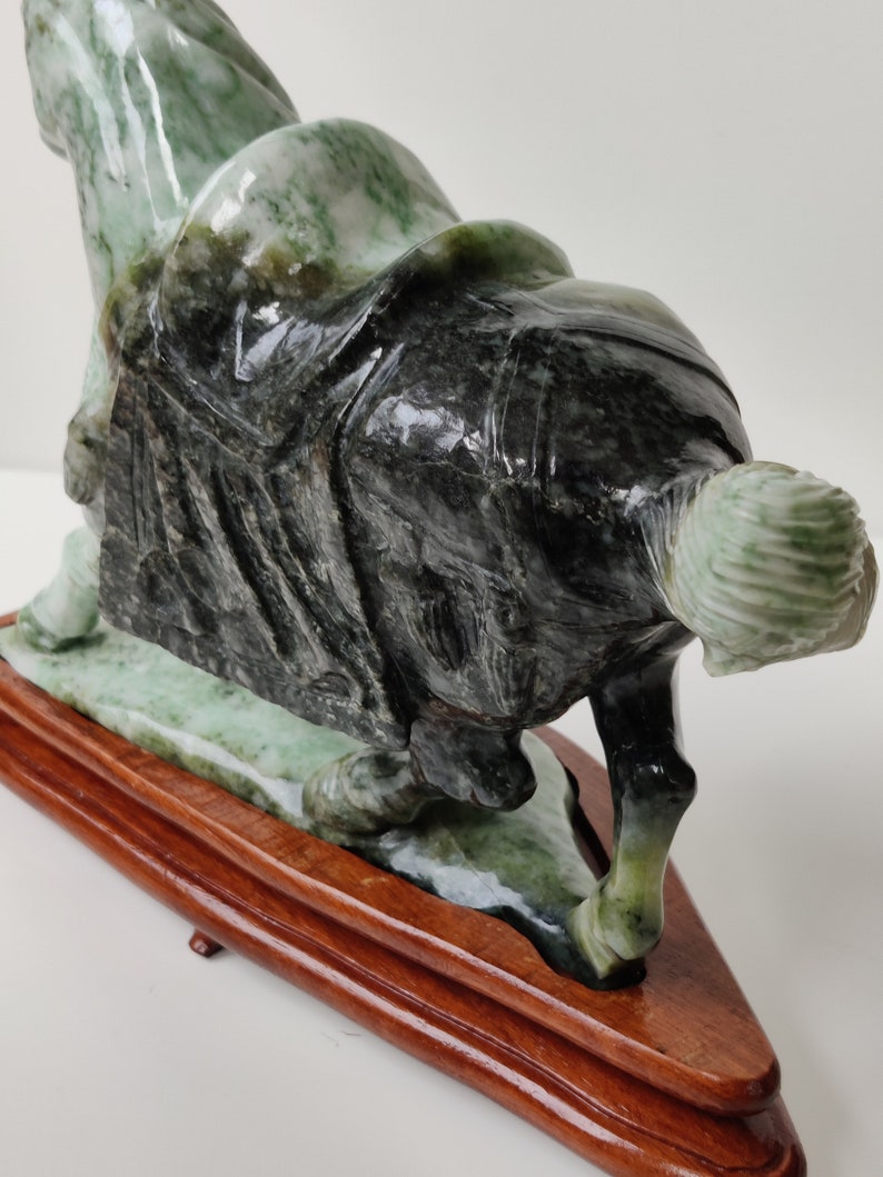 May include: A green and black jade sculpture of a person kneeling on a wooden base. The sculpture is highly detailed and has a glossy finish.