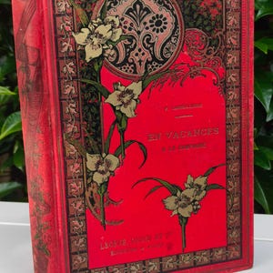 May include: A vintage book with a red cover featuring gold floral illustrations and the title "EN VACANCES A LA CREPINE." The cover has a decorative border and a circular design. The book's spine is also red.