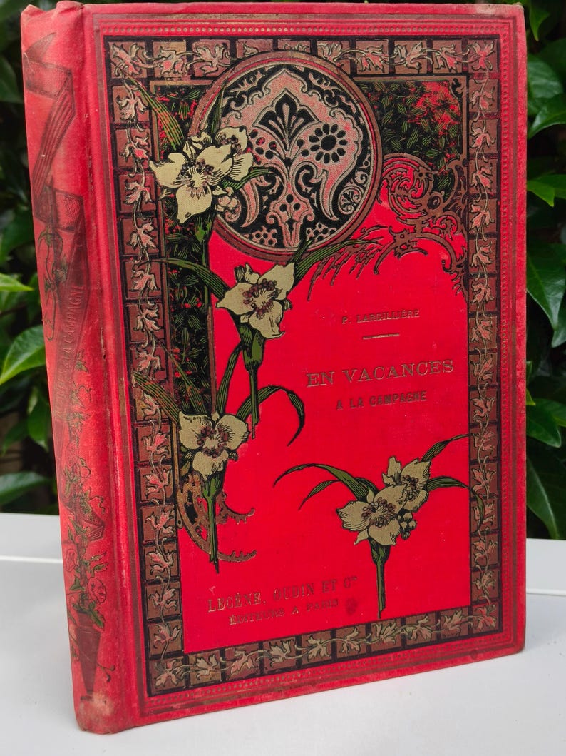May include: A vintage book with a red cover featuring gold floral illustrations and the title "EN VACANCES A LA CREPINE." The cover has a decorative border and a circular design. The book's spine is also red.