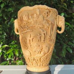 Puede incluir: Un jarrón beige tallado intrincadamente con un relieve detallado que representa un paisaje con figuras y edificios. El jarrón tiene asas con forma de cabeza de elefante y se asienta sobre una base negra. El diseño general es ornamentado y detallado.