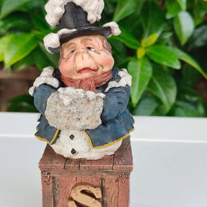 May include: A whimsical figurine of a chef with a rosy face, wearing a black hat with white trim and a blue coat. The chef is standing on a wooden box with a large gold "S" on the front. The figurine has a vintage, handcrafted appearance.