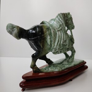 May include: A green jade sculpture of a horse with a rider, mounted on a wooden base. The horse is carved in a detailed style, with a flowing mane and tail. The rider is also carved in detail, with a flowing robe and a sword.