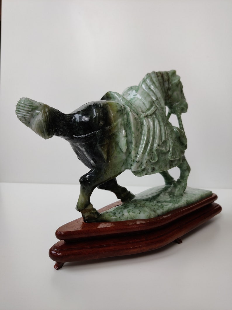 May include: A green jade sculpture of a horse with a rider, mounted on a wooden base. The horse is carved in a detailed style, with a flowing mane and tail. The rider is also carved in detail, with a flowing robe and a sword.
