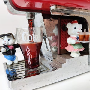 May include: A red and silver Coca-Cola dispenser with a white teddy bear wearing a pink dress and a white teddy bear wearing sunglasses and a blue jacket. The dispenser is pouring a glass of Coca-Cola.