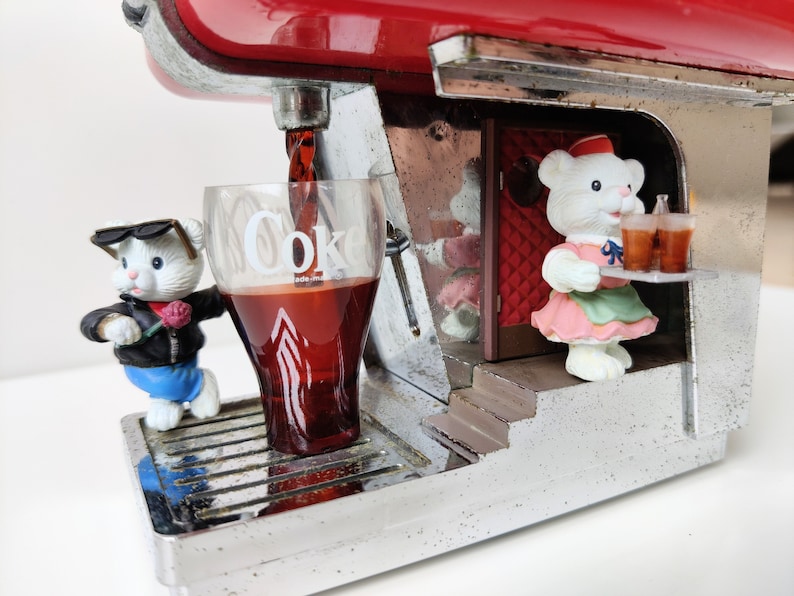 May include: A red and silver Coca-Cola dispenser with a white teddy bear wearing a pink dress and a white teddy bear wearing sunglasses and a blue jacket. The dispenser is pouring a glass of Coca-Cola.
