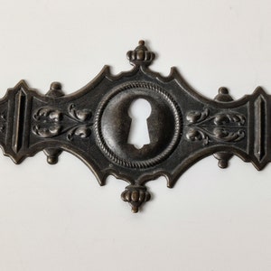 May include: An antique-style keyhole cover in a dark bronze finish. The decorative piece features a central keyhole surrounded by a rope-like border, with floral and scrollwork details. The overall shape is elongated with decorative finials.