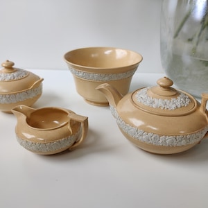 Puede incluir: Un juego de cuatro tazas de té de cerámica de color crema con diseños florales blancos. El juego incluye una tetera, una jarra de crema, un azucarero y un tazón más grande.