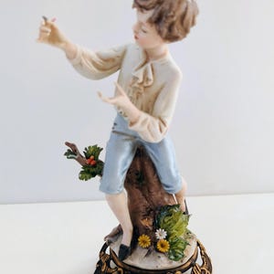 Könnte beinhalten: Eine Porzellanfigur eines Jungen in blauer Hose und weißem Hemd, der auf einem Baumstumpf sitzt und einen Vogel in der Hand hält. Die Figur steht auf einem goldfarbenen Sockel.