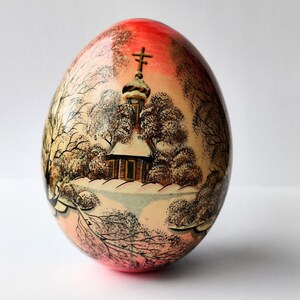 Puede incluir: Un huevo de madera pintado a mano con una escena de invierno. El huevo está pintado con una combinación de colores rojo, blanco y marrón. La escena representa una pequeña iglesia con una cruz en la parte superior, rodeada de árboles y nieve.