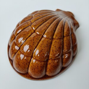 May include: A brown ceramic shell-shaped mold with a speckled glaze. The mold is designed to create a cake or dessert in the shape of a seashell.