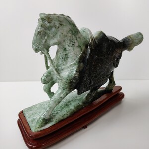 May include: A green jade sculpture of a horse with a rider on a wooden base. The horse is standing on its hind legs and the rider is holding the reins.