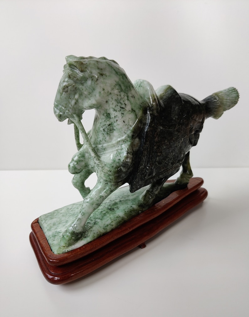 May include: A green jade sculpture of a horse with a rider on a wooden base. The horse is standing on its hind legs and the rider is holding the reins.