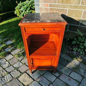 May include: A wooden nightstand with a marble top. The nightstand has a drawer with a round knob, an open shelf, and a small cabinet door. The wood has a reddish-brown finish. The marble top has a dark gray and white pattern.