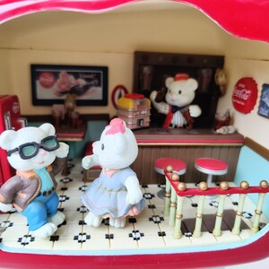 May include: A miniature red Coca-Cola diner playset with a white tiled floor, a counter with stools, and four figurines. The figurines include a bear wearing a red and white striped shirt, a bear wearing a blue jacket and sunglasses, a bear wearing a blue dress, and a bear wearing a red and white striped shirt. The diner has a Coca-Cola sign and a Coca-Cola logo on the wall.