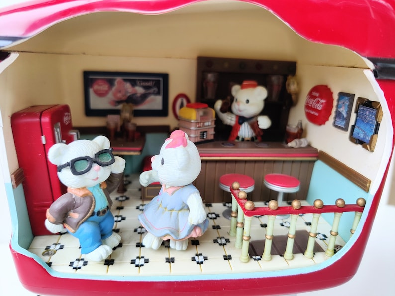 May include: A miniature red Coca-Cola diner playset with a white tiled floor, a counter with stools, and four figurines. The figurines include a bear wearing a red and white striped shirt, a bear wearing a blue jacket and sunglasses, a bear wearing a blue dress, and a bear wearing a red and white striped shirt. The diner has a Coca-Cola sign and a Coca-Cola logo on the wall.