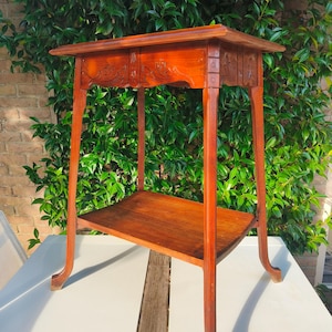 Puede incluir: Una mesa auxiliar de madera vintage con un acabado marrón rojizo intenso. La mesa tiene una tapa rectangular con detalles florales tallados y un estante inferior. Las patas son elegantemente curvadas. La mesa mide unos 76 cm de altura.