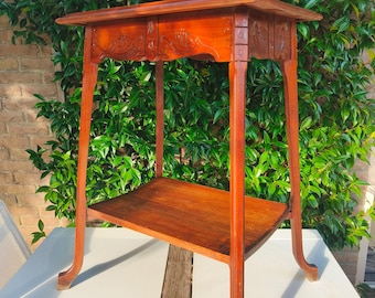 Puede incluir: Una mesa auxiliar de madera vintage con un acabado marrón rojizo intenso. La mesa tiene una tapa rectangular con detalles florales tallados y un estante inferior. Las patas son elegantemente curvadas. La mesa mide unos 76 cm de altura.