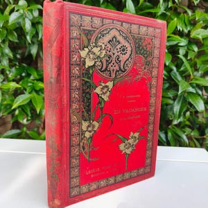 May include: A vintage red book with gold floral and geometric designs. The cover features the title "EN VACANCES" and the author's name. The book is standing upright, showcasing its ornate cover against a green leafy background.