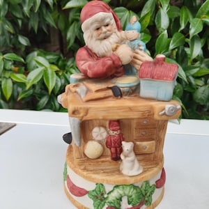 Puede incluir: Una figura navideña de cerámica que representa a Papá Noel en un banco de trabajo, fabricando un juguete. La figura tiene una combinación de colores rojo y blanco, con juguetes adicionales y una base de corona. El fondo es follaje verde.