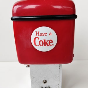May include: Red Coca-Cola vending machine with a white circle that says "Have a Coke" in red letters. The machine has a silver base with two small holes.