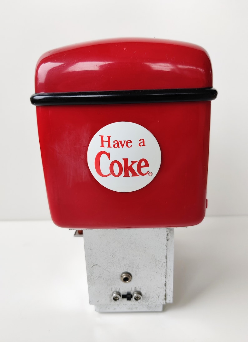 May include: Red Coca-Cola vending machine with a white circle that says "Have a Coke" in red letters. The machine has a silver base with two small holes.