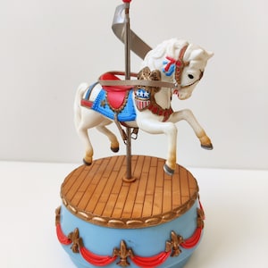 May include: A white carousel horse with a red, white, and blue saddle and a gold trim. The horse is on a blue and gold base with red ribbons. The base has a wooden floor.