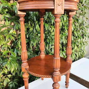 Puede incluir: Una mesa auxiliar redonda de madera de dos niveles con tallas intrincadas en las patas y la parte superior. La mesa está hecha de madera oscura y tiene un acabado natural.