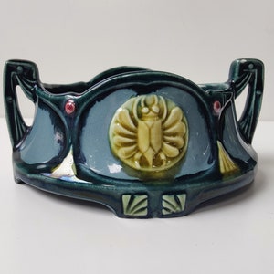 Może przedstawiać: Zielono-niebieska ceramiczna doniczka z dwoma uchwytami i ozdobnym wzorem chrząszcza w środku. Doniczka ma falisty brzeg i wypukły wzór wokół krawędzi.