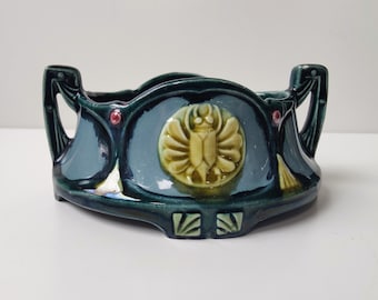 Peut inclure: Un pot en céramique vert et bleu avec deux poignées et un motif de scarabée décoratif au centre. Le pot a un bord festonné et un motif en relief autour du bord.