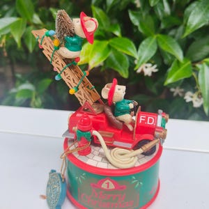 Puede incluir: Una figura musical con temática navideña que presenta dos ratones bomberos. Uno conduce un camión de bomberos rojo, mientras que otro sube una escalera. La base es verde con el texto "Merry Christmas" y un gráfico de casco de bombero.