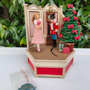 Puede incluir: Figura musical decorativa con una bailarina, un cascanueces y un árbol de Navidad adornado con lazos rojos. El escenario incluye un reloj y regalos, todo dentro de un marco rojo y crema. Se incluye una llave.