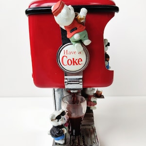 May include: A red Coca-Cola dispenser with a silver base and a glass of cola being poured. The dispenser has a white polar bear figurine climbing on the side and the words "Have a Coke" on a round silver plaque.