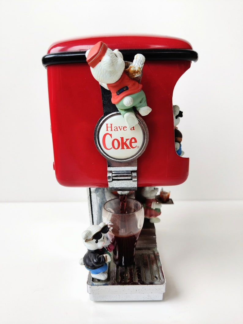 May include: A red Coca-Cola dispenser with a silver base and a glass of cola being poured. The dispenser has a white polar bear figurine climbing on the side and the words "Have a Coke" on a round silver plaque.