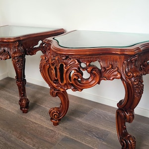 May include: Two ornate, brown wooden console tables with glass tops. The tables feature intricate carvings and curved legs, showcasing a classic, antique design. The tables are placed against a white wall and rest on a wooden floor.