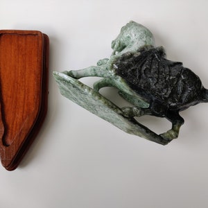 May include: A green and black jade horse sculpture on a wooden stand. The horse is carved in a dynamic pose with its head turned to the side. The stand is made of dark wood and has a simple design.
