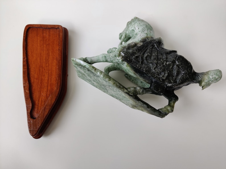 May include: A green and black jade horse sculpture on a wooden stand. The horse is carved in a dynamic pose with its head turned to the side. The stand is made of dark wood and has a simple design.