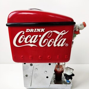 May include: Red Coca-Cola dispenser with a silver base and a polar bear figurine drinking from a glass of Coca-Cola. The dispenser has the text "DRINK Coca-Cola ICE COLD" on the front.