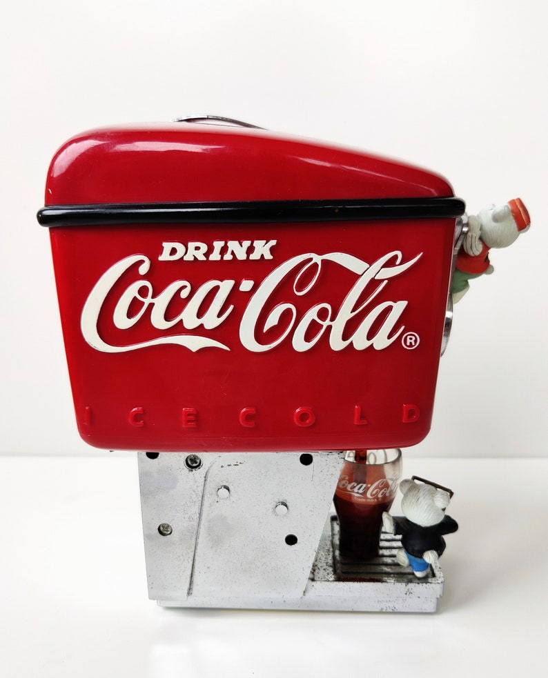 May include: Red Coca-Cola dispenser with a silver base and a polar bear figurine drinking from a glass of Coca-Cola. The dispenser has the text "DRINK Coca-Cola ICE COLD" on the front.