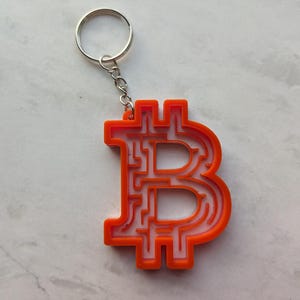 May include: Orange plastic keychain shaped like a Bitcoin symbol with a silver key ring. The Bitcoin symbol is a maze design.