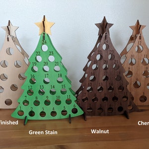May include: Four wooden Christmas tree ornaments in different finishes: unfinished, green stain, walnut, and cherry. Each tree has 24 numbered circles for a Christmas countdown.