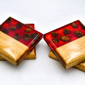 May include: Set of four square coasters made of wood with a red epoxy resin top. The resin has a design of pine cones embedded in it.