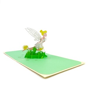 Puede incluir: Una tarjeta pop-up 3D con un hada con alas blancas y un vestido blanco sentada en un mont&iacute;culo de hierba verde con flores amarillas.