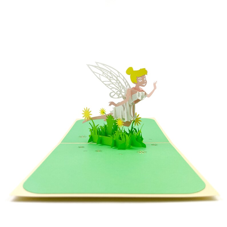 Puede incluir: Una tarjeta pop-up 3D con un hada de pelo rubio y alas blancas, sentada en una cama de hierba verde con flores amarillas.