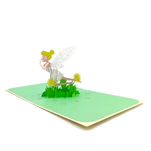 Puede incluir: Una tarjeta pop-up 3D con un hada con alas, un vestido blanco y cabello rubio. El hada est&aacute; sentada en un mont&iacute;culo de hierba verde con flores amarillas.