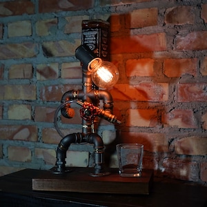 May include: A steampunk-style table lamp made from pipes and fittings, with a light bulb and a bottle of whiskey on top. The lamp is sitting on a wooden base and has a glass shot glass next to it.