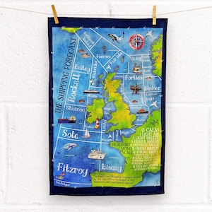 Pode incluir: Um pano de cozinha azul marinho com um mapa das Ilhas Britânicas, indicando as áreas de previsão marítima. O mapa inclui ilustrações de navios, baleias e uma rosa dos ventos. Texto: "The Shipping Forecast" e escala de Beaufort.
