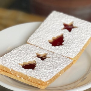 May include: Two square-shaped cookies with a white powdered sugar coating and a red jelly filling in the shape of a star. The cookies are on a white plate.