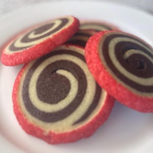May include: Close-up of several spiral cookies on a white plate. The cookies have a red outer edge, a light tan center, and a dark brown spiral design. The cookies are arranged in a slightly overlapping pattern.