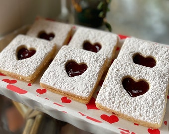 May include: A tray of heart-shaped jam-filled cookies dusted with powdered sugar. The cookies are square with a heart cut out, revealing the red jam filling. The tray has a red and white heart pattern.