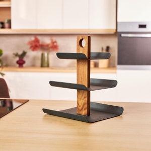 Könnte beinhalten: Ein dreistufiger schwarzer Metallständer mit einem Holzpfosten in der Mitte. Der Ständer hat ein geschwungenes Design und ist perfekt zum Präsentieren von Artikeln wie Snacks, Toilettenartikeln oder Schmuck.
